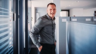 Ein lachender Vertriebsmitarbeiter in moderner Büroumgebung lebt die Vielfalt und Dynamik im Sales-Alltag mit ständig neuen Kundenkontakten, Herausforderungen und Lösungsansätzen. 