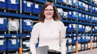 Projektmanagerin mit Laptop in der Hand steht vor einem Hochregallager mit blauen Behältern – symbolisiert Verantwortung, Organisationstalent und strategisches Arbeiten im Bereich Projektmanagement innerhalb logistischer Prozesse.