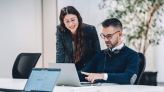 Zwei Mitarbeitende tauschen sich vor einem Laptop aus, was wirkungsvolles Teamwork mit direktem Einfluss auf die Effizienz und Nachhaltigkeit globaler Lieferketten in der Intralogistik symbolisiert. 