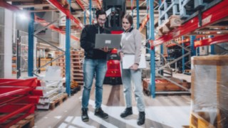 Zwei Mitarbeitende stehen in einem automatisierten Hochregallager vor einem Linde-Fahrzeug und besprechen technische Inhalte am Laptop, was die Arbeit an modernen Automatisierungslösungen in einem innovativen, technologiegetriebenen Umfeld symbolisiert. 