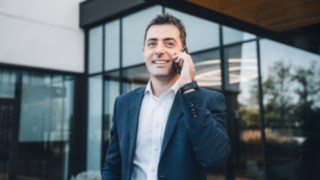 Ein Mitarbeiter im Business-Outfit telefoniert lächelnd vor dem Bürogebäude und ist engagiert im Bereich Inbetriebnahme bei Linde. 