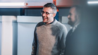 Kollege im grauen Strickpullover lacht herzlich im Gespräch mit einem Teammitglied, während er bei Linde im Bereich Inbetriebnahme arbeitet. 
