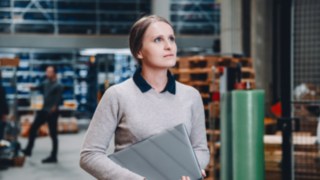 Eine technikaffine Mitarbeiterin mit Laptop unter dem Arm blickt konzentriert in einer modernen Lagerumgebung als Teil des Inbetriebnahme-Teams von Linde. 