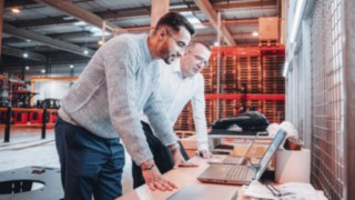 Zwei Kollegen analysieren Projektpläne an Laptops in einer modernen Lagerumgebung und demonstrieren kreative Zusammenarbeit bei KION Automation. 