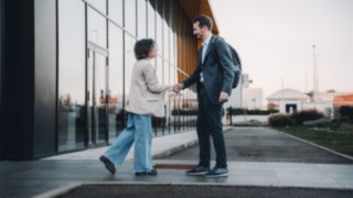 Zwei internationale Kolleg:innen begrüßen sich vor dem Firmengebäude und symbolisieren das globale Arbeiten bei KION Automation. 