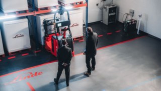 Zwei Fachkräfte begutachten ein autonomes Linde-Fahrzeug in einer modern ausgestatteten Lagerumgebung, was den Einsatz hochentwickelter Automatisierungslösungen und die aktive Mitgestaltung technologischer Innovation repräsentiert. 