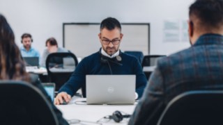 Ein Mitarbeiter mit Headset arbeitet konzentriert an einem Laptop in einem modernen, kollaborativen Büro, was kontinuierliche berufliche Weiterentwicklung durch gezielte Schulungen und individuelle Förderprogramme symbolisiert. 