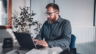 Ein Mitarbeiter arbeitet konzentriert am Laptop in einem hellen Büro mit natürlichem Lichteinfall, was flexible Arbeitsbedingungen symbolisiert, die eine ausgewogene Work-Life-Balance und produktives Arbeiten im Homeoffice oder Büro ermöglichen. 