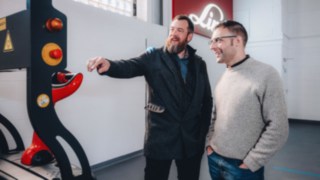 Zwei Kollegen führen ein Gespräch vor einem automatisierten Linde-Fahrzeug in einer hellen Werkshalle, was für offene Kommunikation, technisches Teamwork und eine Arbeitskultur steht, die von Zusammenarbeit und gegenseitiger Unterstützung geprägt ist. 