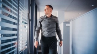 Ein Mitarbeiter geht selbstbewusst durch einen modernen Büroflur mit Glaswänden, was persönliche Verantwortung und einen aktiven Beitrag zur Effizienz und Nachhaltigkeit globaler Lieferketten symbolisiert. 