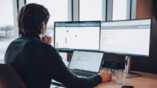 Ein KION Automation-Mitarbeiter arbeitet an technischen Zeichnungen und Automatisierungsplänen an einem Schreibtisch mit drei Bildschirmen, was den Einsatz modernster Technologien in der Intralogistik symbolisiert.