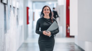 Eine selbstbewusste Mitarbeiterin mit Laptop in der Hand geht lächelnd durch einen modernen Büroflur und steht für engagiertes Arbeiten an Projekten, die globale Lieferketten effizienter und nachhaltiger gestalten. 