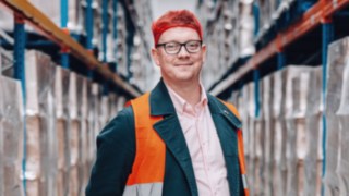 Ein Mitarbeiter aus dem Bereich Solution Design bei KION Automation, mit Warnweste und roter Haarnetzkappe, steht professionell und freundlich in einem Hochregallager.