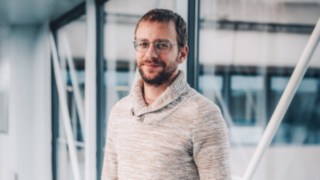 Ein KION Automation-Mitarbeiter aus dem Solution Design lächelt in die Kamera, aufgenommen vor moderner Büroarchitektur mit Glasfassade. 