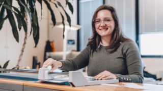 Eine Mitarbeiterin aus dem Bereich Solution Design bei KION Automation lächelt freundlich, während sie an einem Schreibtisch mit einem Schneidegerät in einem modernen Büro arbeitet. 