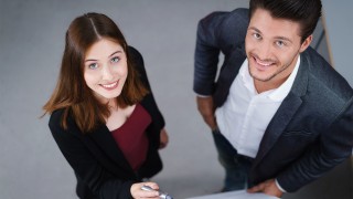 eine Frau und ein Mann, stehen nebeneinander und lächeln freundlich nach oben zur Kamera. Die Frau hält einen Stift.